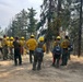 A large crew of wildland firefighters is gathered on a dirt road or clearing in a forested area, appearing to receive a briefing or safety talk. All personnel are wearing standard wildland firefighting gear including yellow Nomex shirts, green pants, and yellow hard hats. The setting appears to be an active or recent fire area based on the dry conditions and some scorched vegetation visible in the background.