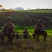 A group of military personnel in full combat gear are positioned across a grassy, hilly terrain during what appears to be a training exercise or tactical operation. The soldiers are armed with assault rifles and wearing camouflage uniforms, helmets, and tactical vests with face paint applied. The setting appears to be a tropical or subtropical environment, likely at dawn or dusk based on the lighting conditions.