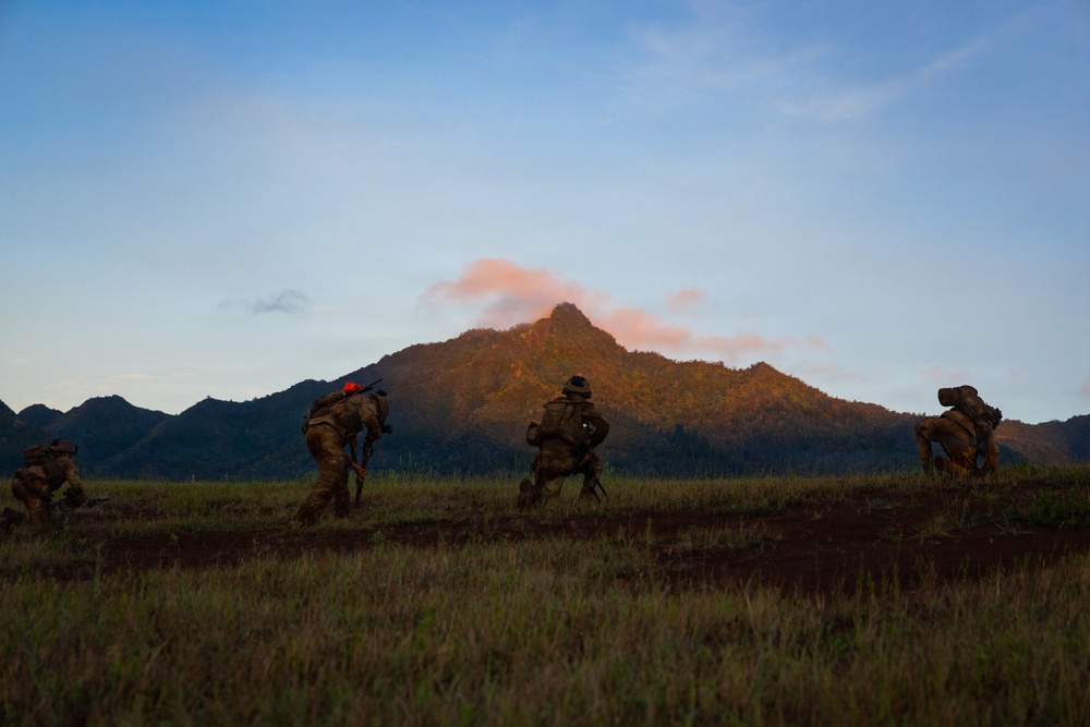 The image shows multiple military personnel in camouflage uniforms moving tactically across an open grassy field at dusk or dawn. They are silhouetted against a dramatic backdrop of a peaked mountain illuminated by warm golden-pink light. The scene appears to depict a military training exercise or field operation.