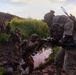 A group of military personnel in full combat gear are navigating through a muddy, vegetated terrain during what appears to be a training exercise at dusk or dawn. One soldier in the foreground is assisting another across a small water obstacle, while a column of additional soldiers follows behind. The lush green landscape with tall grass and a hillside in the background suggests a tropical or subtropical environment.