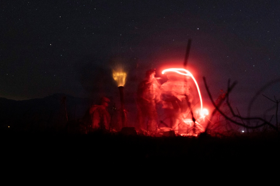 Marines fire weapons in the dark under a starry sky illuminated by red and orange lights.
