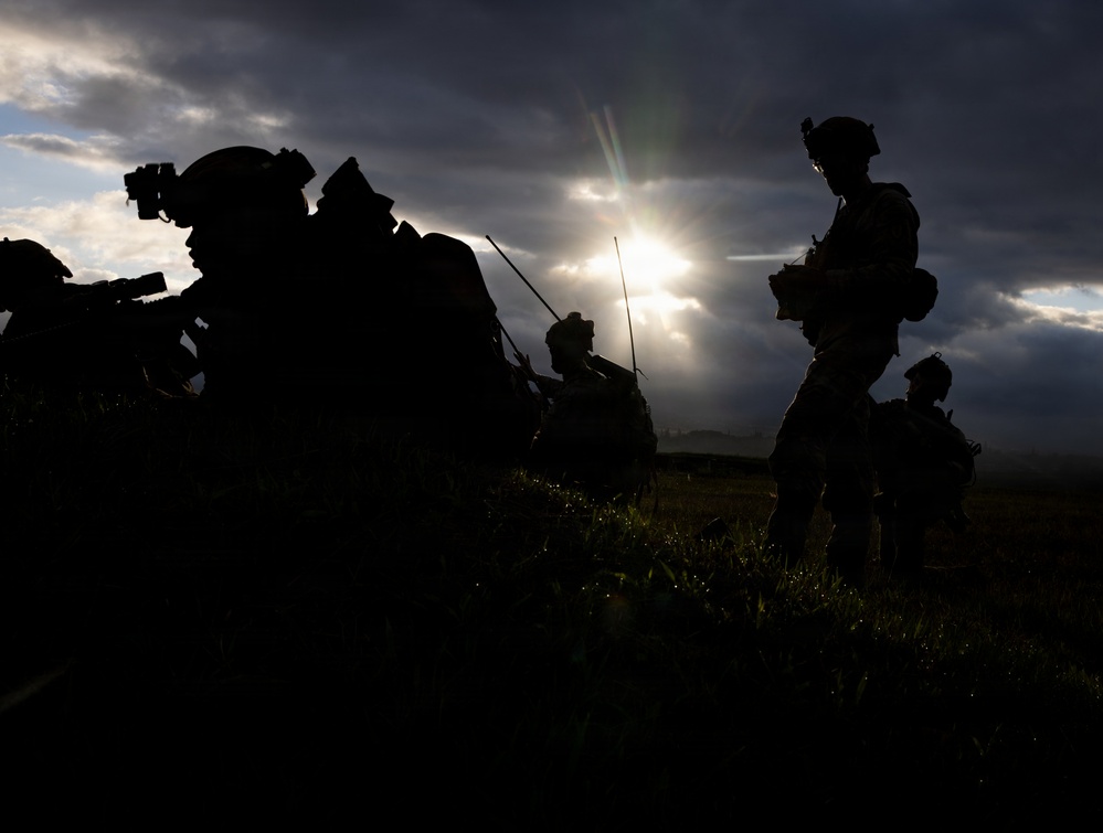 A dramatic silhouette photograph showing multiple military personnel in full tactical gear positioned in an open field. The subjects are backlit by a partially obscured sun breaking through dark storm clouds, creating stark black silhouettes. The image captures what appears to be a tactical pause or briefing during a military operation or exercise.