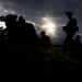A dramatic silhouette photograph showing multiple military personnel in full tactical gear positioned in an open field. The subjects are backlit by a partially obscured sun breaking through dark storm clouds, creating stark black silhouettes. The image captures what appears to be a tactical pause or briefing during a military operation or exercise.