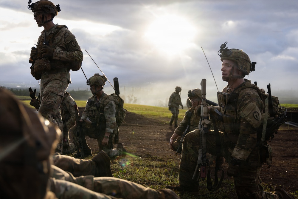 A group of military personnel in full combat gear are assembled on an open grassy field, appearing to be during a training exercise or operational briefing. The soldiers are equipped with rifles, tactical vests, helmets with night-vision mounts, and radio antennas. The scene is backlit by dramatic sunlight breaking through overcast clouds, creating a high-contrast atmospheric setting.