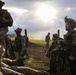 A group of military personnel in full combat gear are assembled on an open grassy field, appearing to be during a training exercise or operational briefing. The soldiers are equipped with rifles, tactical vests, helmets with night-vision mounts, and radio antennas. The scene is backlit by dramatic sunlight breaking through overcast clouds, creating a high-contrast atmospheric setting.