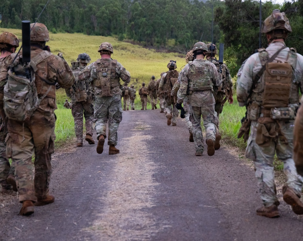 A column of military personnel in full combat gear marches down a narrow paved road through a green, hilly landscape. The soldiers are photographed from behind, moving away from the camera in a loose formation. The setting appears to be a training exercise or patrol in a rural, subtropical environment.