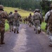 A column of military personnel in full combat gear marches down a narrow paved road through a green, hilly landscape. The soldiers are photographed from behind, moving away from the camera in a loose formation. The setting appears to be a training exercise or patrol in a rural, subtropical environment.