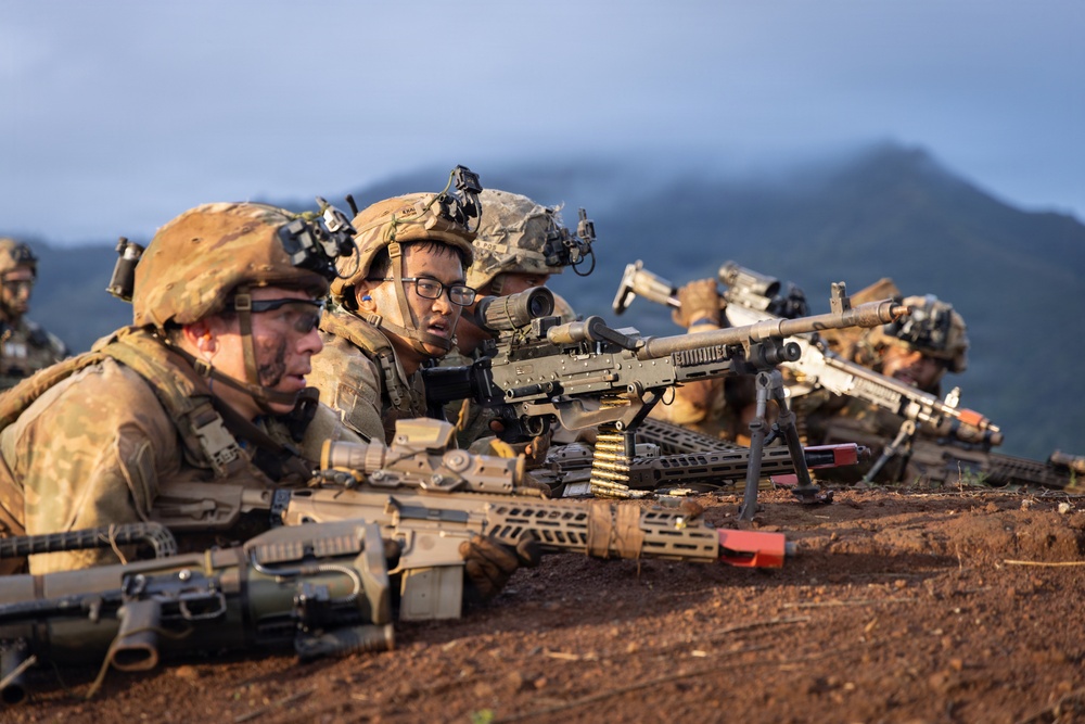 A group of soldiers in full combat gear are positioned prone on a dirt berm, aiming weapons downrange in what appears to be a military training exercise. Multiple personnel are equipped with helmets featuring night-vision mounts, camouflage uniforms, and various weapon systems including machine guns on bipods. The mountainous, lush background and red soil suggest a tropical or subtropical training environment.