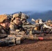 A group of soldiers in full combat gear are positioned prone on a dirt berm, aiming weapons downrange in what appears to be a military training exercise. Multiple personnel are equipped with helmets featuring night-vision mounts, camouflage uniforms, and various weapon systems including machine guns on bipods. The mountainous, lush background and red soil suggest a tropical or subtropical training environment.