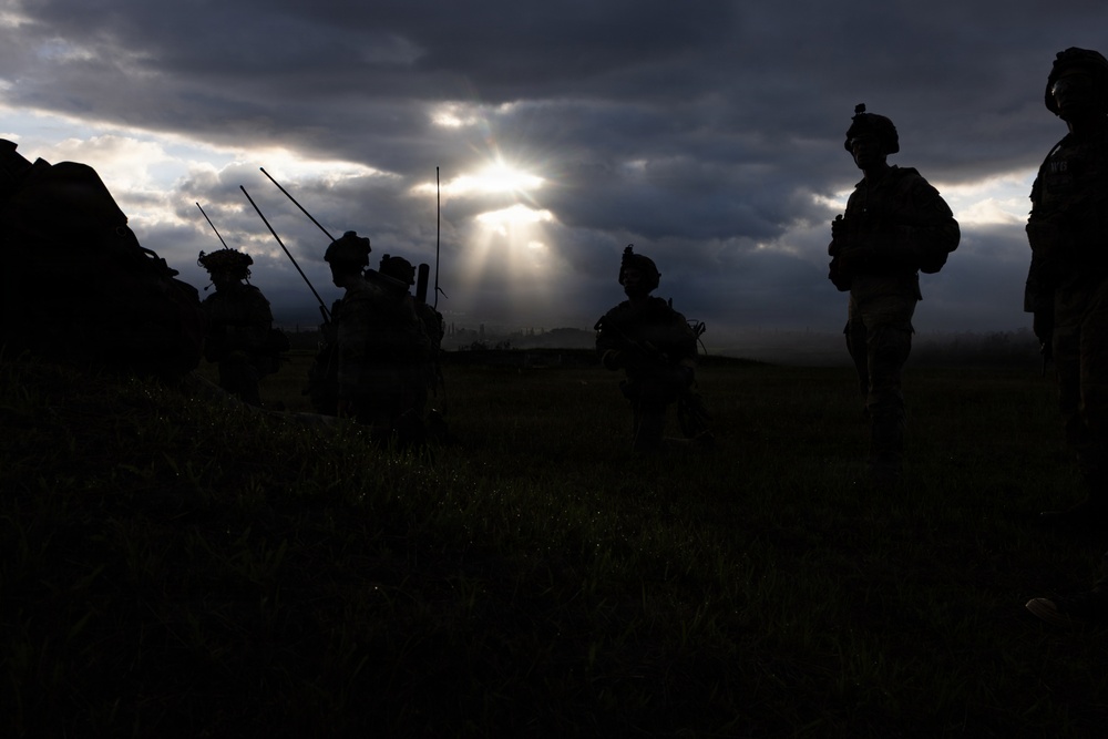 A dramatic silhouette photograph showing a group of military personnel in full combat gear standing in an open field. The image is backlit by rays of sunlight breaking through dark, stormy clouds, creating a stark contrast between the dark figures and the illuminated sky. The soldiers appear to be in a formation or briefing posture.