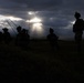 A dramatic silhouette photograph showing a group of military personnel in full combat gear standing in an open field. The image is backlit by rays of sunlight breaking through dark, stormy clouds, creating a stark contrast between the dark figures and the illuminated sky. The soldiers appear to be in a formation or briefing posture.