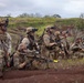 A group of heavily armed military personnel in full combat gear are positioned along a dirt embankment in a green, hilly terrain. The soldiers appear to be in a tactical observation posture, scanning their surroundings during what appears to be a training exercise or field operation. Multiple soldiers are equipped with night vision devices, body armor, and various weapons systems.