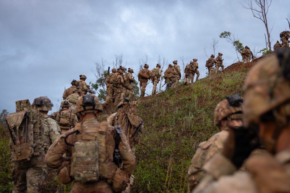 A large group of soldiers in full combat gear and camouflage uniforms are ascending a steep grassy hillside in a column formation. The terrain appears to be a tropical or subtropical environment with sparse trees and red-brown exposed soil. This appears to be a military training exercise or tactical movement operation.