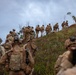 A large group of soldiers in full combat gear and camouflage uniforms are ascending a steep grassy hillside in a column formation. The terrain appears to be a tropical or subtropical environment with sparse trees and red-brown exposed soil. This appears to be a military training exercise or tactical movement operation.