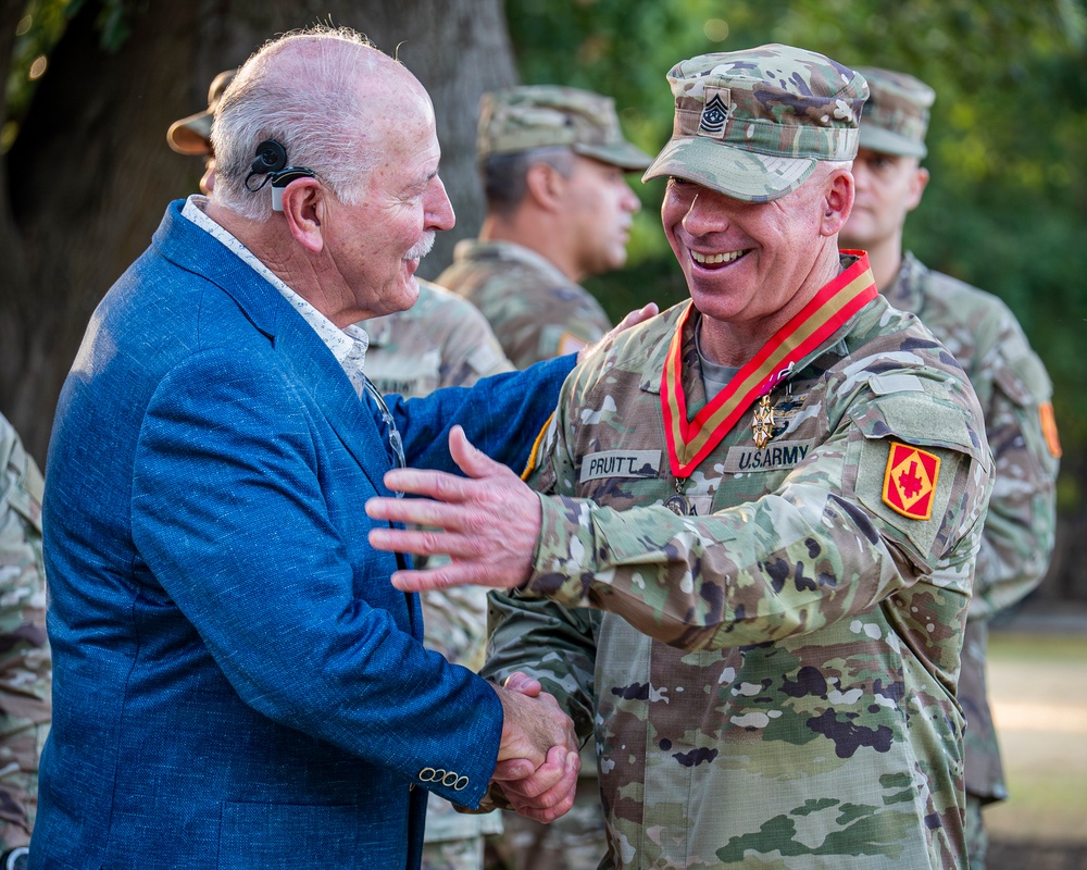 A U.S. Army soldier named Pruitt, wearing a red-ribboned medal/decoration around his neck, shakes hands and shares a warm moment with an elderly civilian man in a blue suit. The soldier is in OCP (Operational Camouflage Pattern) uniform with a Command Sergeant Major rank insignia on his cap and a unit patch on his sleeve. The setting appears to be an outdoor military ceremony with other uniformed soldiers visible in the background.
