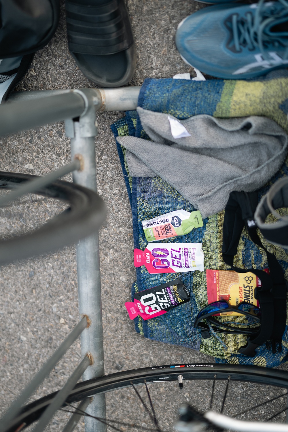 A triathlon or endurance sports transition area setup showing energy nutrition products laid out on a towel next to a bicycle. Multiple energy gel brands are visible alongside cycling/running gear. The scene suggests an athlete preparing for or recovering from a multi-sport event.