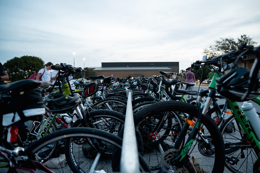 A large collection of bicycles are densely packed into an outdoor bike rack area, likely at a sporting or community event. The photo is taken at ground level during dusk or early evening, giving the scene a low-light, atmospheric quality. Several people can be seen in the background near what appears to be a building or venue.