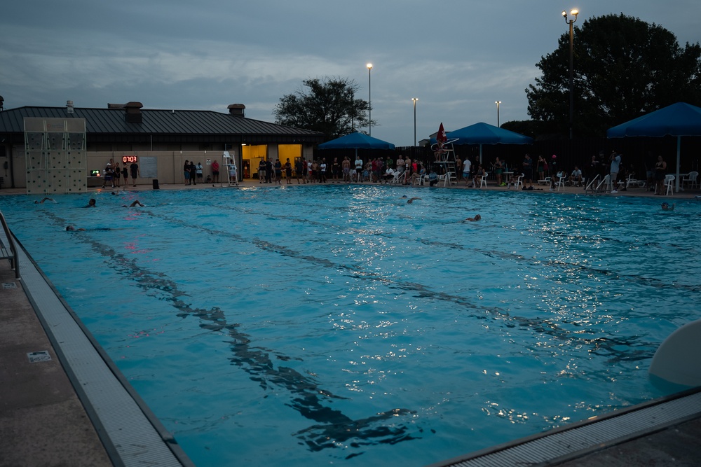 An outdoor swimming pool competition or practice session photographed at dusk or early evening under overcast skies. Multiple swimmers are visible in the lanes while a crowd of spectators lines the far side of the pool. A digital scoreboard/timer is visible on the left wall of the pool facility building.