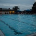 An outdoor swimming pool competition or practice session photographed at dusk or early evening under overcast skies. Multiple swimmers are visible in the lanes while a crowd of spectators lines the far side of the pool. A digital scoreboard/timer is visible on the left wall of the pool facility building.