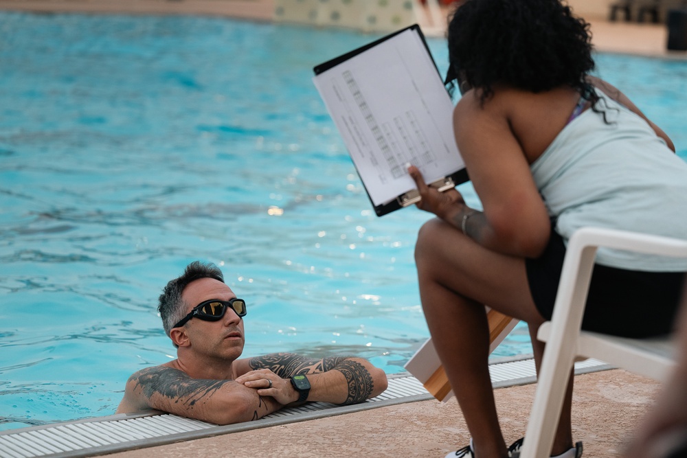A tattooed man wearing sunglasses and a smartwatch is in a swimming pool, resting his arms on the pool edge while looking up at a woman seated poolside. The woman, wearing a light blue top and black shorts, is holding a clipboard with documents and appears to be reviewing or discussing information with him. The setting appears to be an outdoor pool area with tiled surroundings.