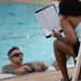 A tattooed man wearing sunglasses and a smartwatch is in a swimming pool, resting his arms on the pool edge while looking up at a woman seated poolside. The woman, wearing a light blue top and black shorts, is holding a clipboard with documents and appears to be reviewing or discussing information with him. The setting appears to be an outdoor pool area with tiled surroundings.