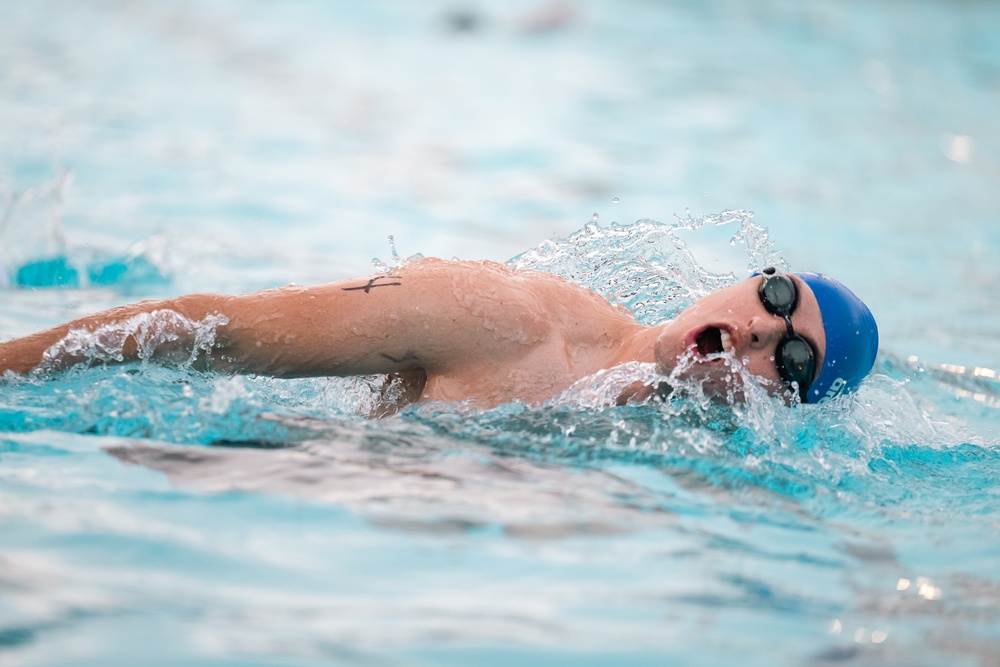 A competitive swimmer is captured mid-stroke performing the freestyle (front crawl) technique in a swimming pool. The swimmer is wearing a blue swim cap and goggles, with their head turned to the side for a breath. Water splashes dynamically around the athlete, suggesting high-speed movement.
