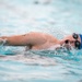 A competitive swimmer is captured mid-stroke performing the freestyle (front crawl) technique in a swimming pool. The swimmer is wearing a blue swim cap and goggles, with their head turned to the side for a breath. Water splashes dynamically around the athlete, suggesting high-speed movement.