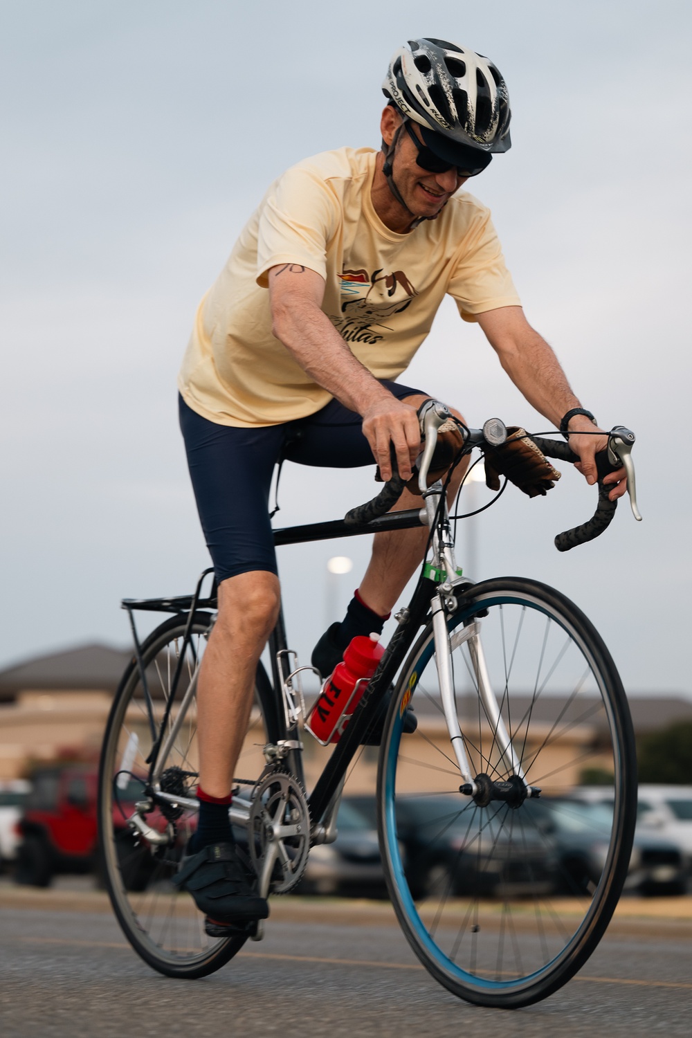 A man is riding a road bicycle in what appears to be a parking lot. He is wearing a yellow t-shirt, navy blue cycling shorts, black shoes with red socks, sunglasses, and a black-and-white helmet. He appears to be smiling and looking downward while pedaling.