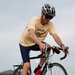 A man is riding a road bicycle in what appears to be a parking lot. He is wearing a yellow t-shirt, navy blue cycling shorts, black shoes with red socks, sunglasses, and a black-and-white helmet. He appears to be smiling and looking downward while pedaling.