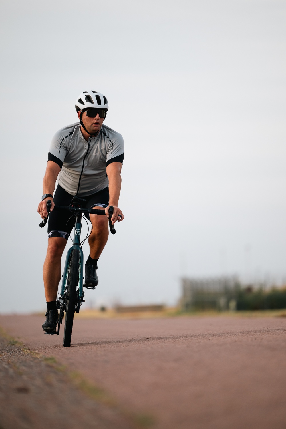 A male cyclist is riding a teal/blue road or gravel bike directly toward the camera on a reddish-brown paved path. He is dressed in full cycling kit including a white vented helmet, sport sunglasses, a gray and black short-sleeve jersey, black bib shorts, and black cycling shoes. The background is a flat, open landscape with blurred trees and an overcast sky.
