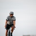 A male cyclist is riding a teal/blue road or gravel bike directly toward the camera on a reddish-brown paved path. He is dressed in full cycling kit including a white vented helmet, sport sunglasses, a gray and black short-sleeve jersey, black bib shorts, and black cycling shoes. The background is a flat, open landscape with blurred trees and an overcast sky.
