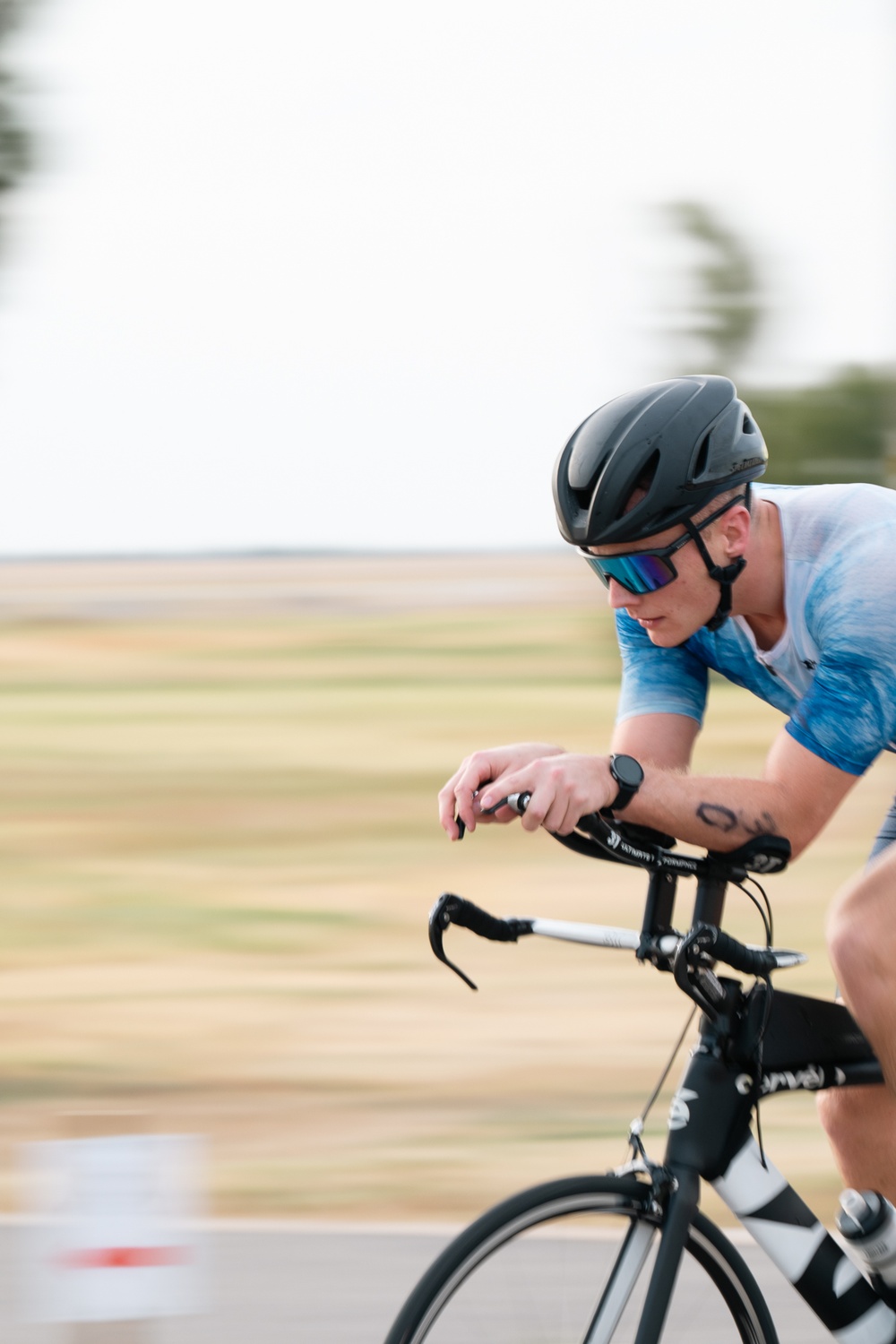 A cyclist is captured in motion on a paved road, leaning forward in an aerodynamic position. The rider is equipped with professional cycling gear including a black helmet, blue mirrored sunglasses, and a blue/white jersey. The background is blurred due to motion, suggesting high speed, with flat open terrain visible.