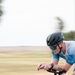 A cyclist is captured in motion on a paved road, leaning forward in an aerodynamic position. The rider is equipped with professional cycling gear including a black helmet, blue mirrored sunglasses, and a blue/white jersey. The background is blurred due to motion, suggesting high speed, with flat open terrain visible.