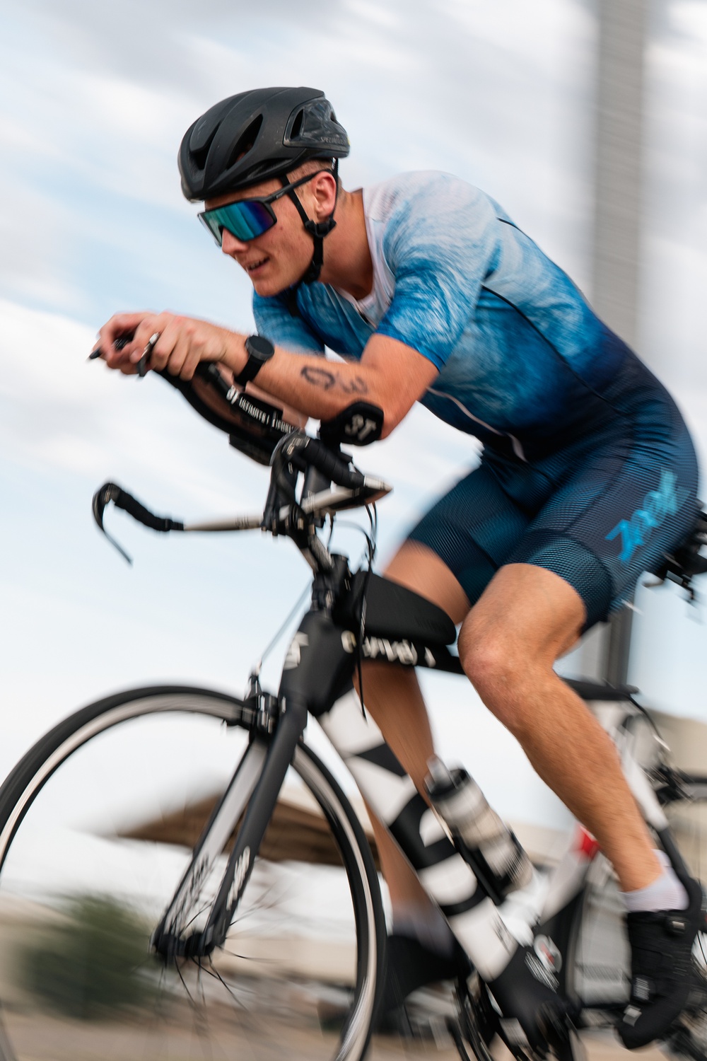 A male cyclist is captured mid-ride in a dynamic, motion-blurred action shot. He is riding an aerodynamic road/triathlon bike in an aggressive forward-leaning position. The image conveys high speed and athletic intensity through deliberate panning blur technique.