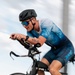 A male cyclist is captured mid-ride in a dynamic, motion-blurred action shot. He is riding an aerodynamic road/triathlon bike in an aggressive forward-leaning position. The image conveys high speed and athletic intensity through deliberate panning blur technique.