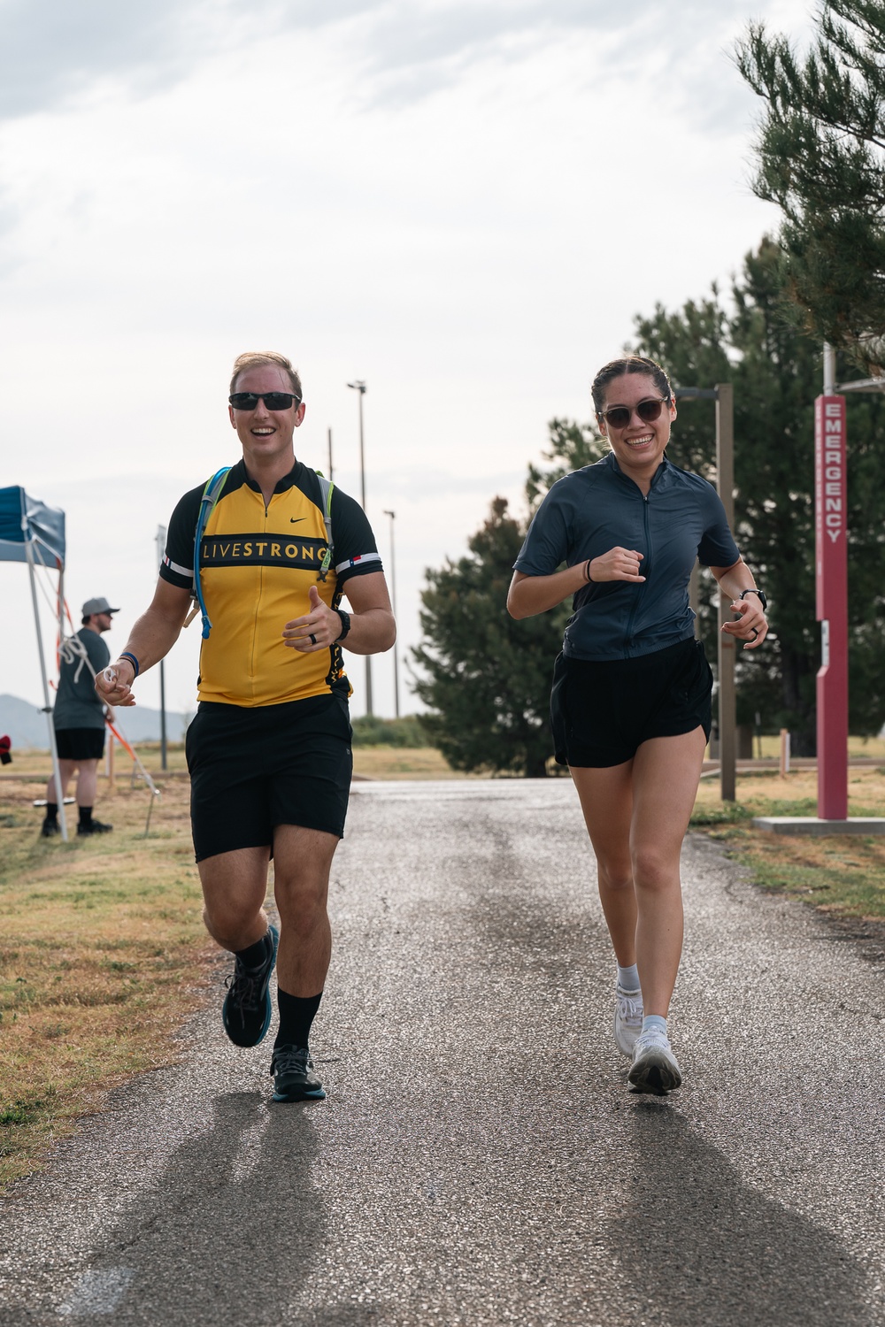 Two people are running together on a paved path, both smiling and appearing to enjoy themselves. The man on the left is wearing a yellow and black 'LIVESTRONG' cycling jersey with a hydration pack, while the woman on the right wears a gray athletic top and black shorts. The setting appears to be an outdoor recreational or athletic facility, possibly a military base given the 'EMERGENCY' post visible in the background.
