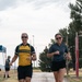 Two people are running together on a paved path, both smiling and appearing to enjoy themselves. The man on the left is wearing a yellow and black 'LIVESTRONG' cycling jersey with a hydration pack, while the woman on the right wears a gray athletic top and black shorts. The setting appears to be an outdoor recreational or athletic facility, possibly a military base given the 'EMERGENCY' post visible in the background.