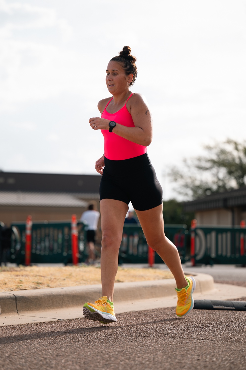 A woman is running outdoors on a paved surface, wearing athletic clothing including a neon pink tank top, black shorts, and yellow running shoes. She has her dark hair in a bun and is wearing a black sports watch on her left wrist. The background shows a fenced area with orange bollards and other people visible in the distance.