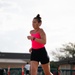 A woman is running outdoors on a paved surface, wearing athletic clothing including a neon pink tank top, black shorts, and yellow running shoes. She has her dark hair in a bun and is wearing a black sports watch on her left wrist. The background shows a fenced area with orange bollards and other people visible in the distance.