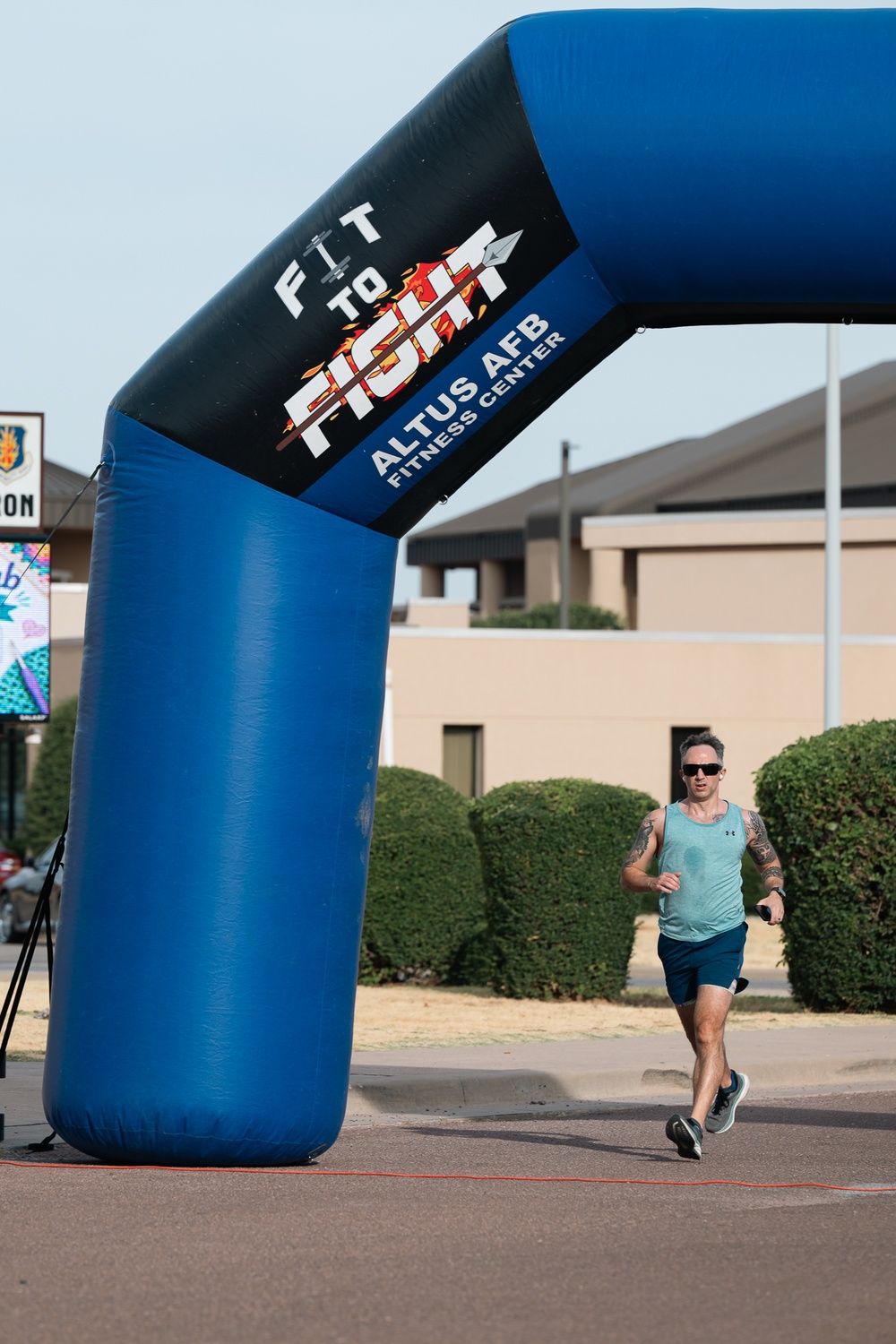 A male runner passes under a large inflatable arch at what appears to be a fitness run event at Altus Air Force Base. The arch is branded with 'Fit to Fight' and 'Altus AFB Fitness Center' logos. The event appears to be taking place on a paved road on the base during daylight hours.