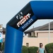 A male runner passes under a large inflatable arch at what appears to be a fitness run event at Altus Air Force Base. The arch is branded with 'Fit to Fight' and 'Altus AFB Fitness Center' logos. The event appears to be taking place on a paved road on the base during daylight hours.