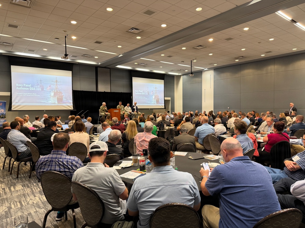 A large professional conference or symposium is taking place in a modern convention center ballroom. The event features a 'Navy Panel Audience Q&amp;A' session, as displayed on two large projection screens showing naval vessels. Several panelists, some in military uniforms, are seated at the front of the room addressing a packed audience of approximately 100-150 attendees.