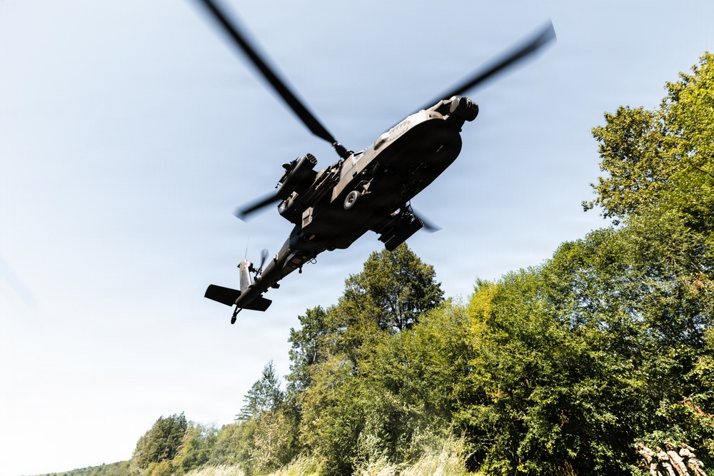 A military attack helicopter, consistent with an AH-64 Apache, is photographed from a low ground angle as it flies overhead at low altitude. The image is taken from below looking up, with the helicopter's underside and rotor system clearly visible against a partly cloudy sky. Dense green deciduous trees are visible in the background, suggesting a forested or rural environment.