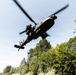 A military attack helicopter, consistent with an AH-64 Apache, is photographed from a low ground angle as it flies overhead at low altitude. The image is taken from below looking up, with the helicopter's underside and rotor system clearly visible against a partly cloudy sky. Dense green deciduous trees are visible in the background, suggesting a forested or rural environment.