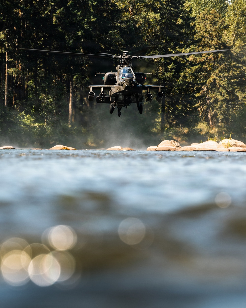 A military attack helicopter, consistent with an AH-64 Apache, is hovering at low altitude over a rocky riverbed surrounded by dense coniferous forest. The photograph is taken from a ground-level perspective at the water's surface, creating a dramatic foreground blur effect with bokeh light reflections. Rotor wash is visibly disturbing dust and debris near the rocks below the aircraft.