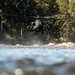 A military attack helicopter, consistent with an AH-64 Apache, is hovering at low altitude over a rocky riverbed surrounded by dense coniferous forest. The photograph is taken from a ground-level perspective at the water's surface, creating a dramatic foreground blur effect with bokeh light reflections. Rotor wash is visibly disturbing dust and debris near the rocks below the aircraft.