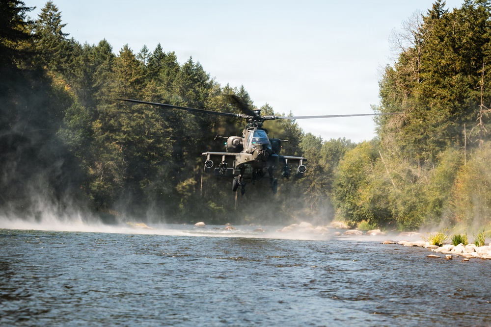 A military attack helicopter, consistent with an AH-64 Apache, is flying at very low altitude over a river in a forested area. The rotor wash is visibly disturbing the water surface and kicking up mist and spray below the aircraft. The surrounding environment consists of dense coniferous and deciduous trees under a partly cloudy sky.