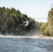 A military attack helicopter, consistent with an AH-64 Apache, is flying at very low altitude over a river in a forested area. The rotor wash is visibly disturbing the water surface and kicking up mist and spray below the aircraft. The surrounding environment consists of dense coniferous and deciduous trees under a partly cloudy sky.