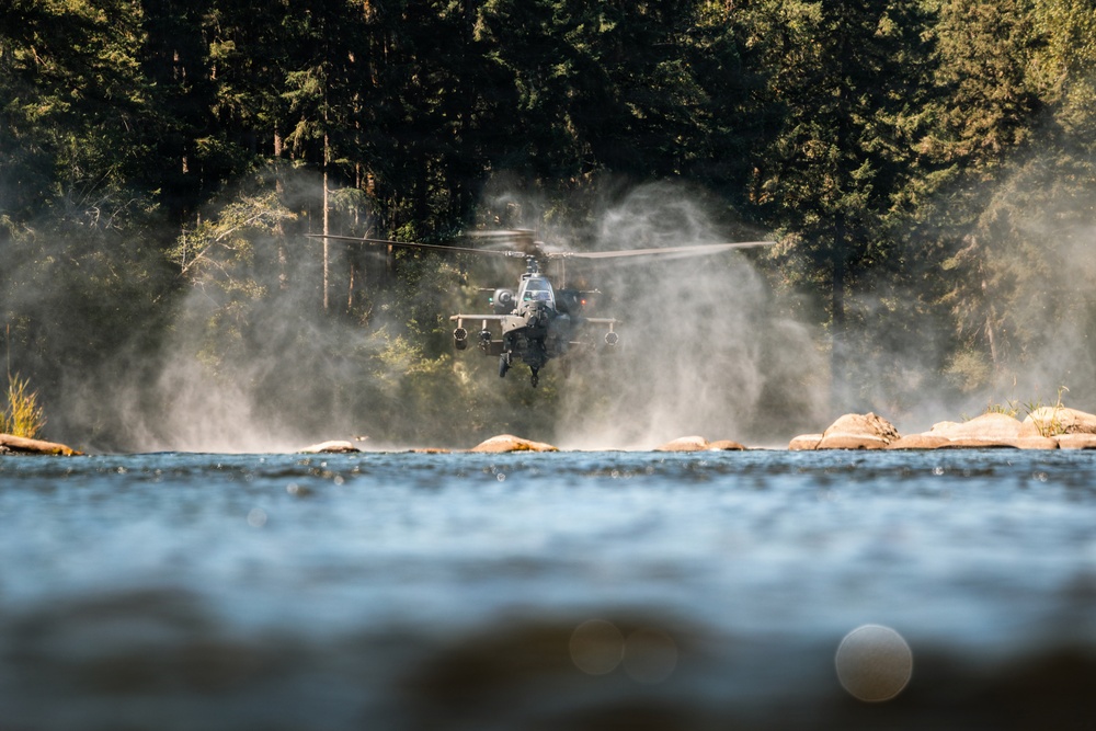 A military attack helicopter, consistent with an AH-64 Apache, is hovering at low altitude over or near a rocky riverbank. The rotor wash is visibly displacing water vapor or mist, creating dramatic spray effects around the aircraft. Dense coniferous forest forms the background, suggesting a Pacific Northwest or similar temperate environment.
