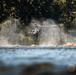 A military attack helicopter, consistent with an AH-64 Apache, is hovering at low altitude over or near a rocky riverbank. The rotor wash is visibly displacing water vapor or mist, creating dramatic spray effects around the aircraft. Dense coniferous forest forms the background, suggesting a Pacific Northwest or similar temperate environment.